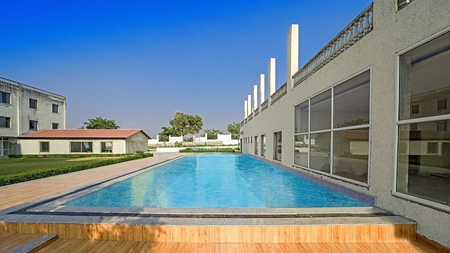 Outdoor swimming pool at Grassfield Riveria Resort, Jaipur, surrounded by modern buildings and a wooden deck area.