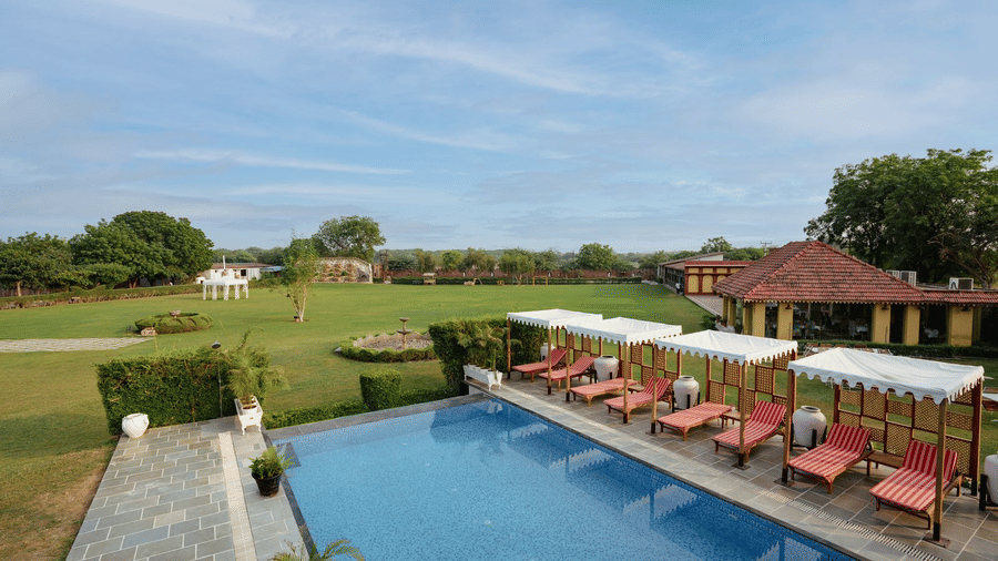 Outdoor swimming pool with sun loungers and shaded cabanas overlooking a large landscaped lawn and heritage-style buildings under a clear blue sky - Surpura Bagh