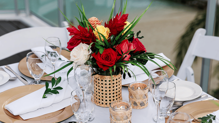 Elegant table setting with a vibrant floral centrepiece of red, yellow, and white roses in a woven vase, surrounded by wine glasses, candles, and neatly arranged place settings.