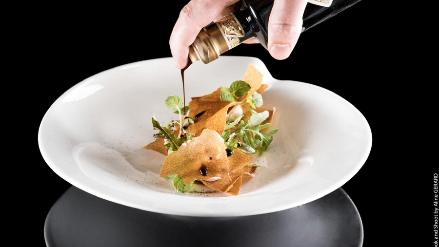Elegant plating in the Taste of France style, showing a hand pouring a dark sauce over a finely prepared dish with crisp garnishes.