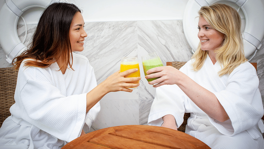 Two women in white robes enjoying fresh green juice in a serene lounge at YO1 Longevity & Health Resorts, nestled in the tranquil Catskills, reflecting relaxation and holistic wellbeing.