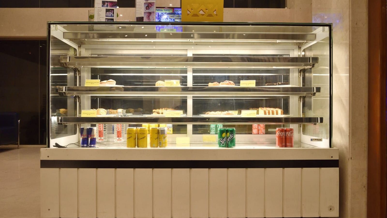 A well-lit refrigerated bakery display case showcasing cakes, pastries, and assorted beverages, located in a modern indoor setting at The Lobby Bake House, The Cunningham Hotel