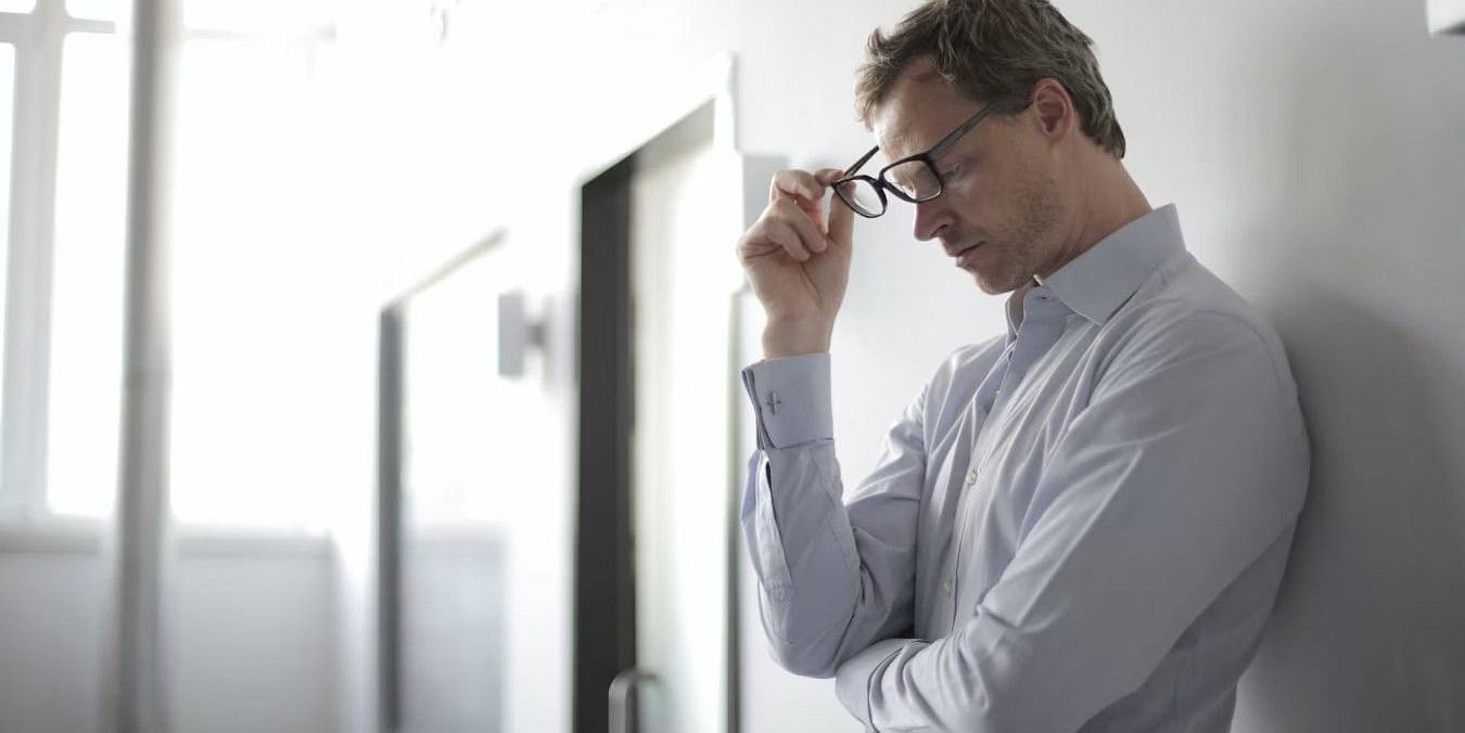 A man in a light blue shirt leans against a white wall, holding his glasses, appearing stressed or deep in thought at YO1 Longevity and Health Resorts