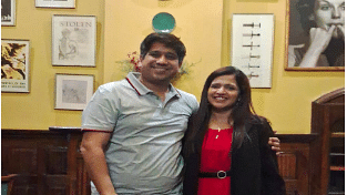 Man and woman posing together and smiling inside a warmly lit room with vintage decor and framed photos.