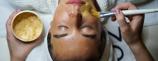 A close-up of a woman receiving a facial treatment, with a paste being applied to her face by a brush.