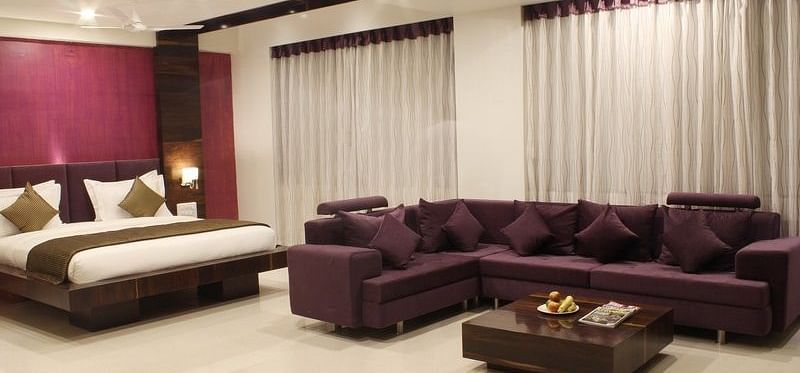 A spacious hotel suite features a king size bed, a comfortable brown sofa set around a coffee table, and large windows with sheer curtains.