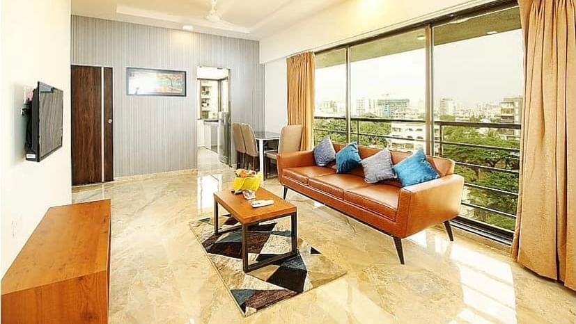 A well-lit living room with a leather sofa, blue cushions, a marble floor, a flat-screen TV on the wall and large windows that offer a view of the city.