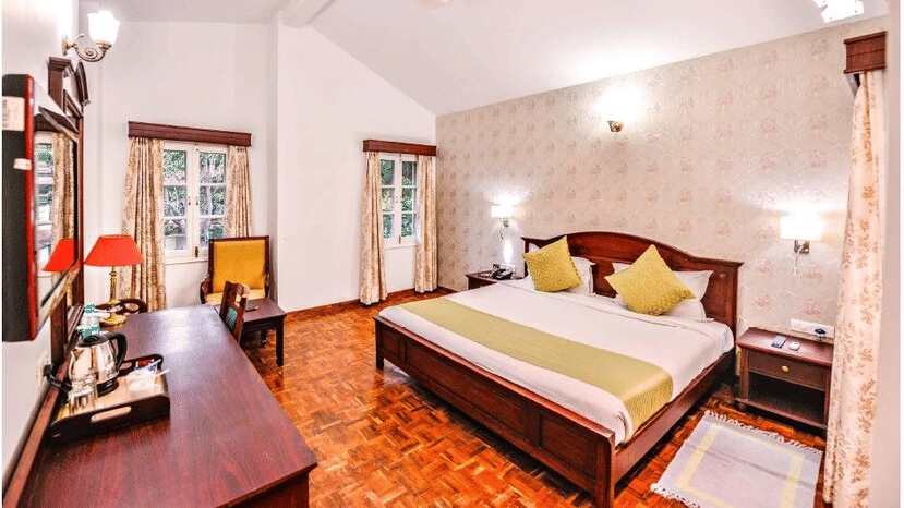 A bedroom with a pitched white ceiling,twin beds with yellow pillows, a work desk, and a chair at Amantra Shilpi Resort, Udaipur.