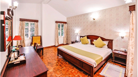 A bedroom with a pitched white ceiling,twin beds with yellow pillows, a work desk, and a chair at Amantra Shilpi Resort, Udaipur.