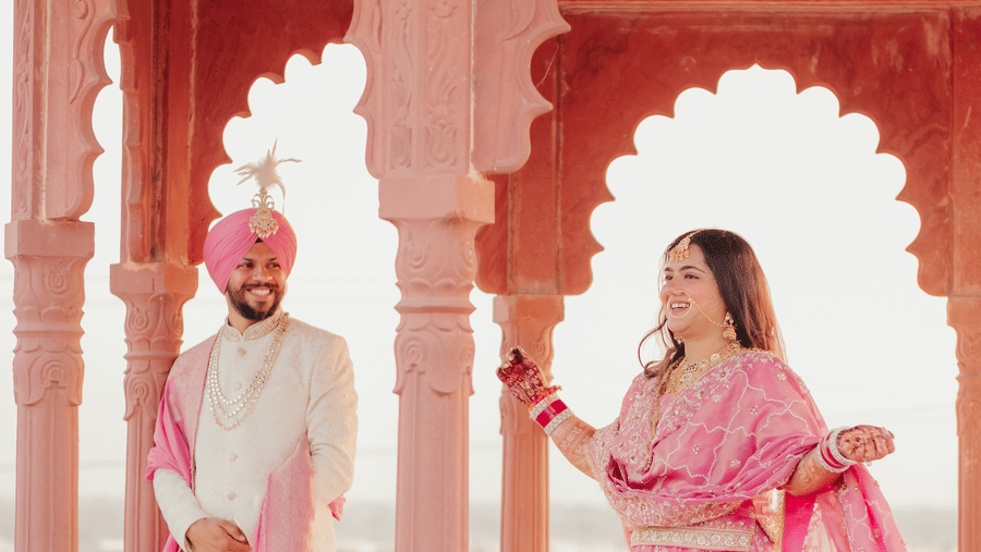 A groom watches his bride walk through an ornate, pink stone traditional arched pavilion at Umaid Palace.