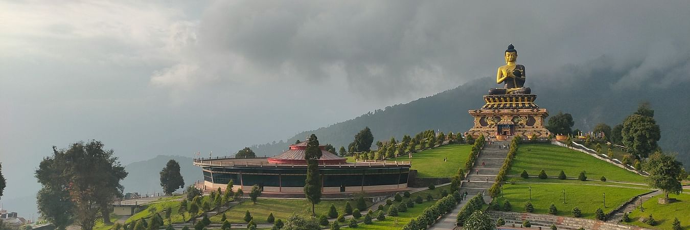 A majestic golden Buddha statue sits atop a hill, surrounded by lush green gardens and mountains under a cloudy sky.