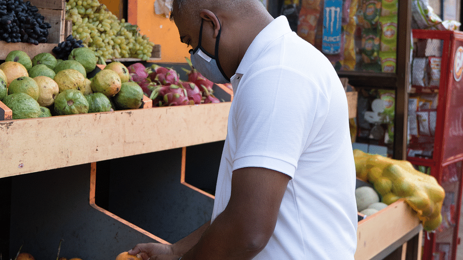 A man wearing a white polo shirt and a dark face mask is selecting fruit, possibly oranges, from a display at an outdoor market stall. Various fruits are visible on shelves behind him.
