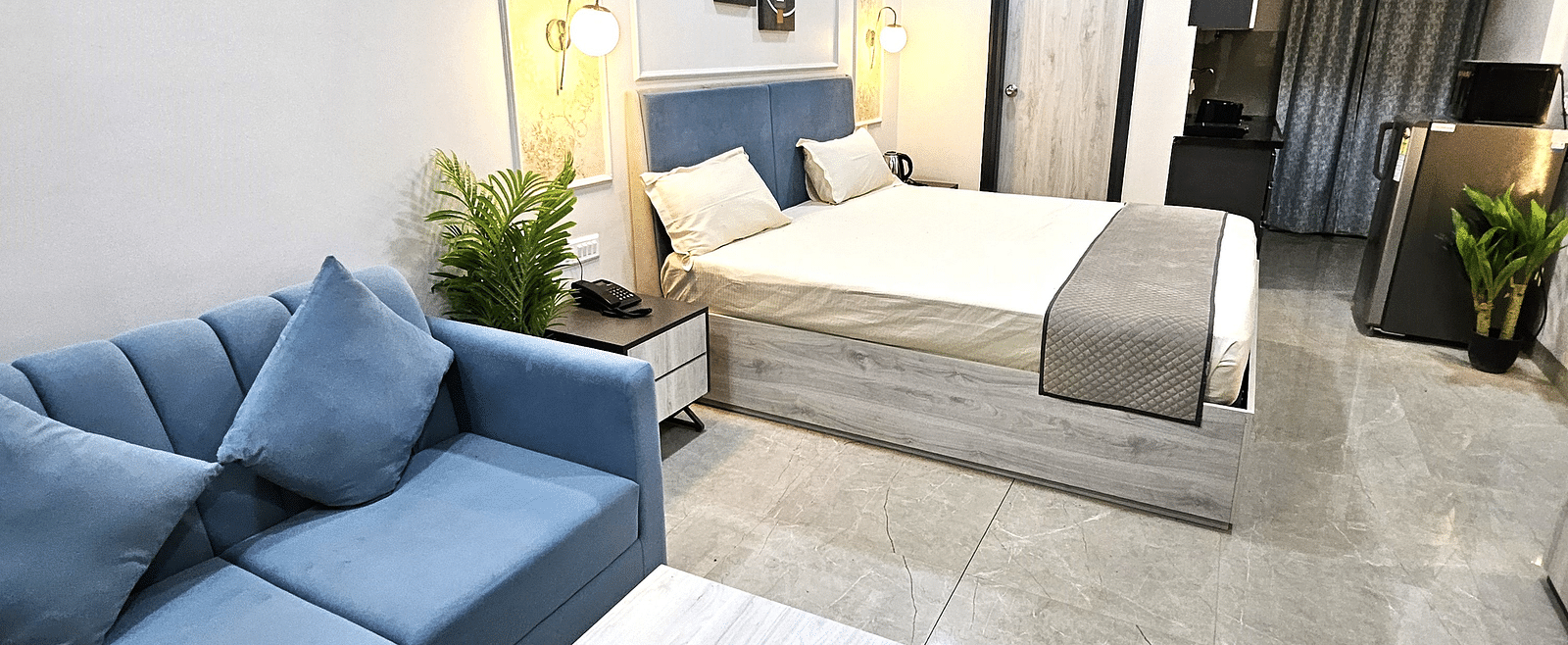 Modern studio room featuring a bed, blue sofa with matching cushions, wooden coffee table, kitchenette with appliances, and warm lighting accents - Urban Suites by BluSalzz, Jaipur