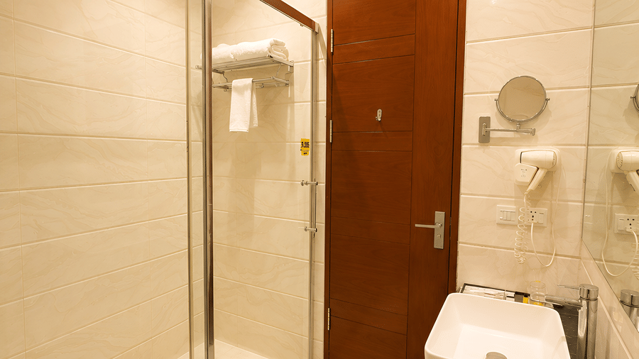 Deluxe room bathroom at Golden Tulip Kukas Jaipur with a glass-enclosed shower, toilet, wooden door, and white tiled walls.