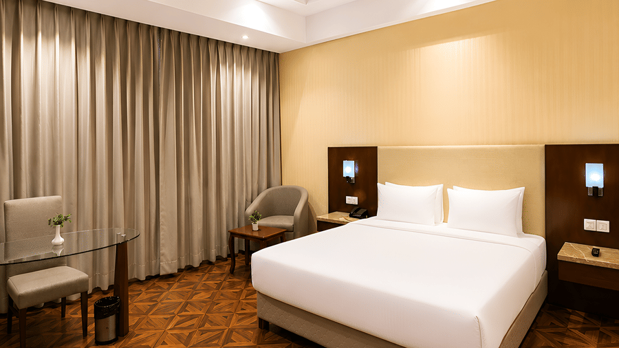Deluxe room at Golden Tulip Kukas Jaipur with a king bed, white bedding, patterned carpet flooring, grey curtains, and soft ambient lighting.