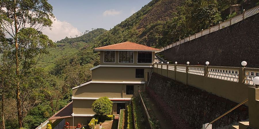 A covered outdoor patio or walkway with a grey metal roof, tiled floor, and a view of trees and a hillside - Abad Copper Castle, Munnar