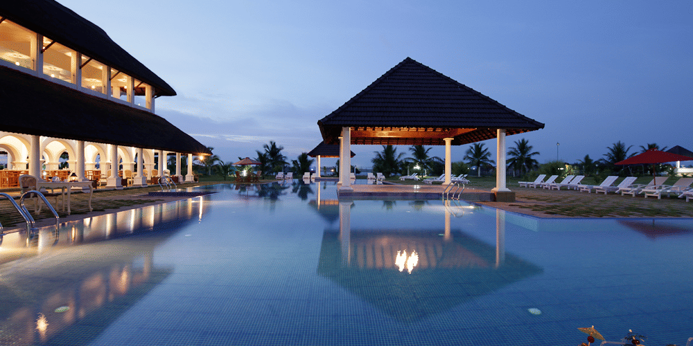 An outdoor swimming pool reflecting a lit pavilion and resort building under a twilight sky with cocktails in the foreground | Le Pondy