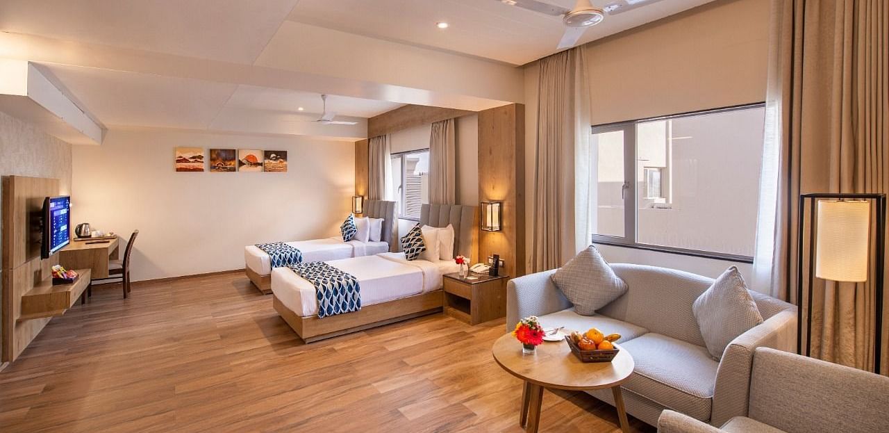 A spacious hotel suite featuring two single beds with blue and white accents, a comfortable seating area with a sofa and armchairs around a small table with fruit, a work desk with a chair, and a television, all on a wooden floor at Lords Inn Motikhavdi