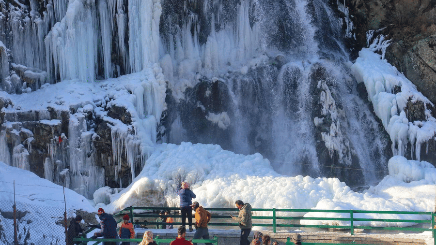 A group of visitors standing at a fenced viewpoint looking at a partially frozen waterfall cliff near Dawat e Drung by FrozenFall Resort.