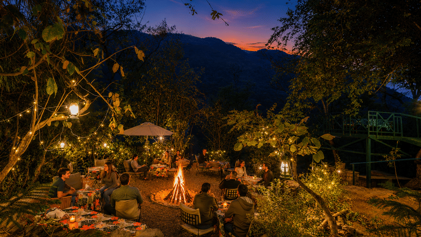 Cosy evening bonfire at Neemrana’s Glasshouse on The Ganges, Rishikesh, with guests relaxing outdoors amidst trees against a sunset over mountains.