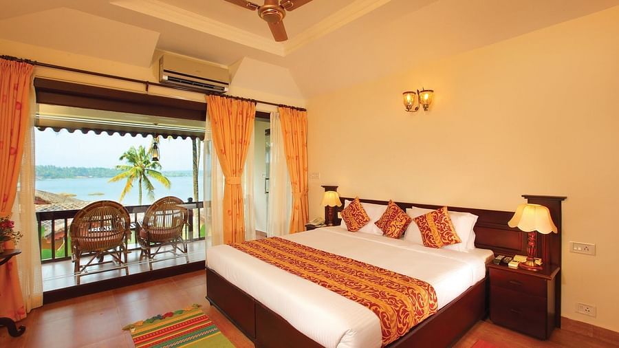 An image of Premium lake View Villa with Private Terrace featuring a double size bed, a bedside table with lamp, and a balcony attached which has two chairs placed- Fragrant Nature Kollam