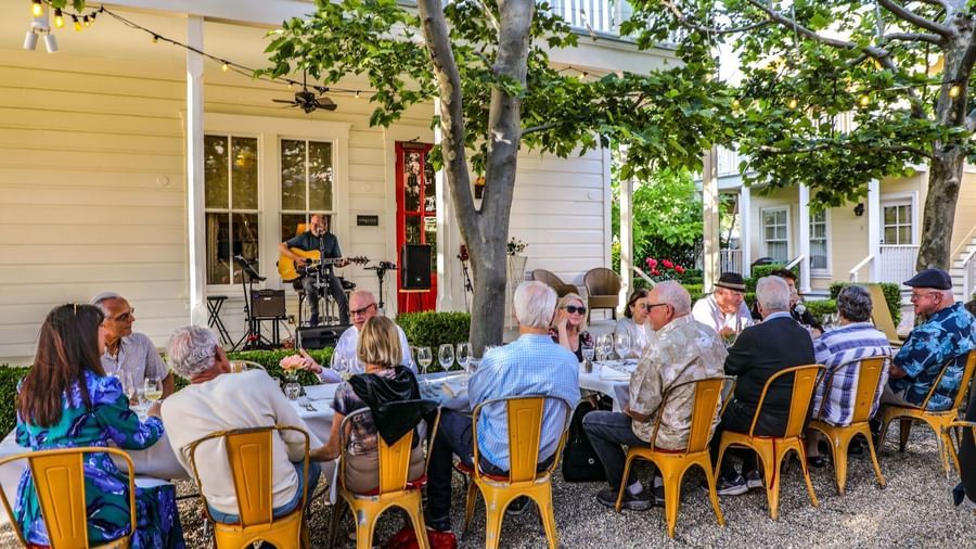 A large group of people seated across two outdoor dining tables with a small white cabin behind at Tallman Hotel.