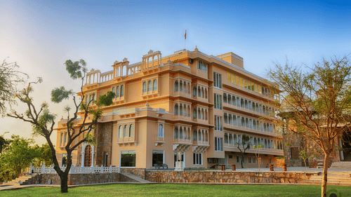Facade of Fateh Niwas in Udaipur featuring a multi storey building and a lawn with trees.