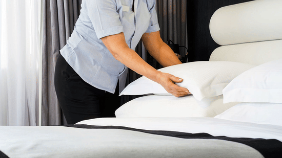 A hotel housekeeper in a striped shirt straightens pillows while making a bed in a guest room.