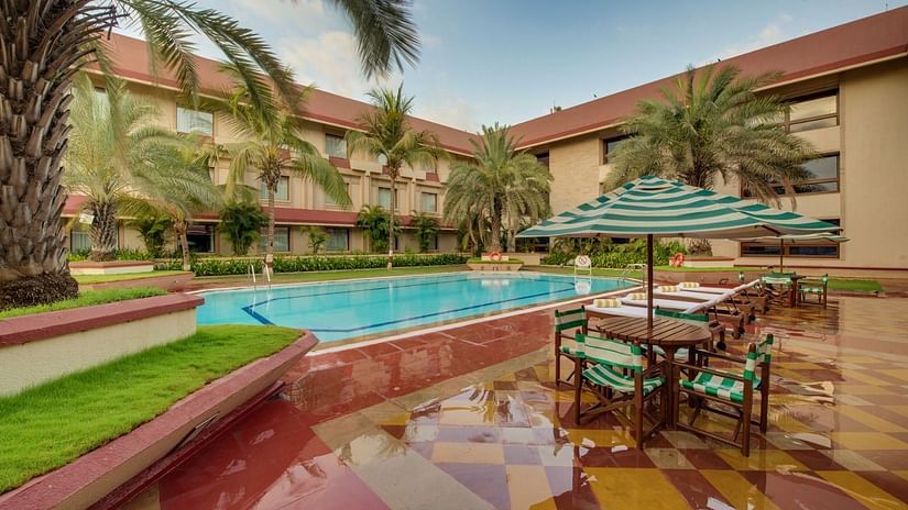 Poolside area at The Ummed Ahmedabad featuring outdoor seating with umbrellas, palm trees, and surrounding hotel buildings under a clear sky.