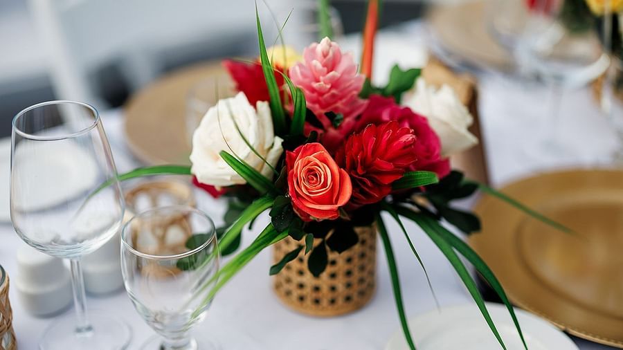 A close-up of a floral centerpiece on a table setting. The centerpiece features red, pink, and white roses in a woven basket. The table is set with plates, glasses, and utensils.