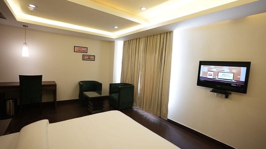 A room with a white bed, a dark table with chairs, and a mounted television on the wall - VITS Gurugram