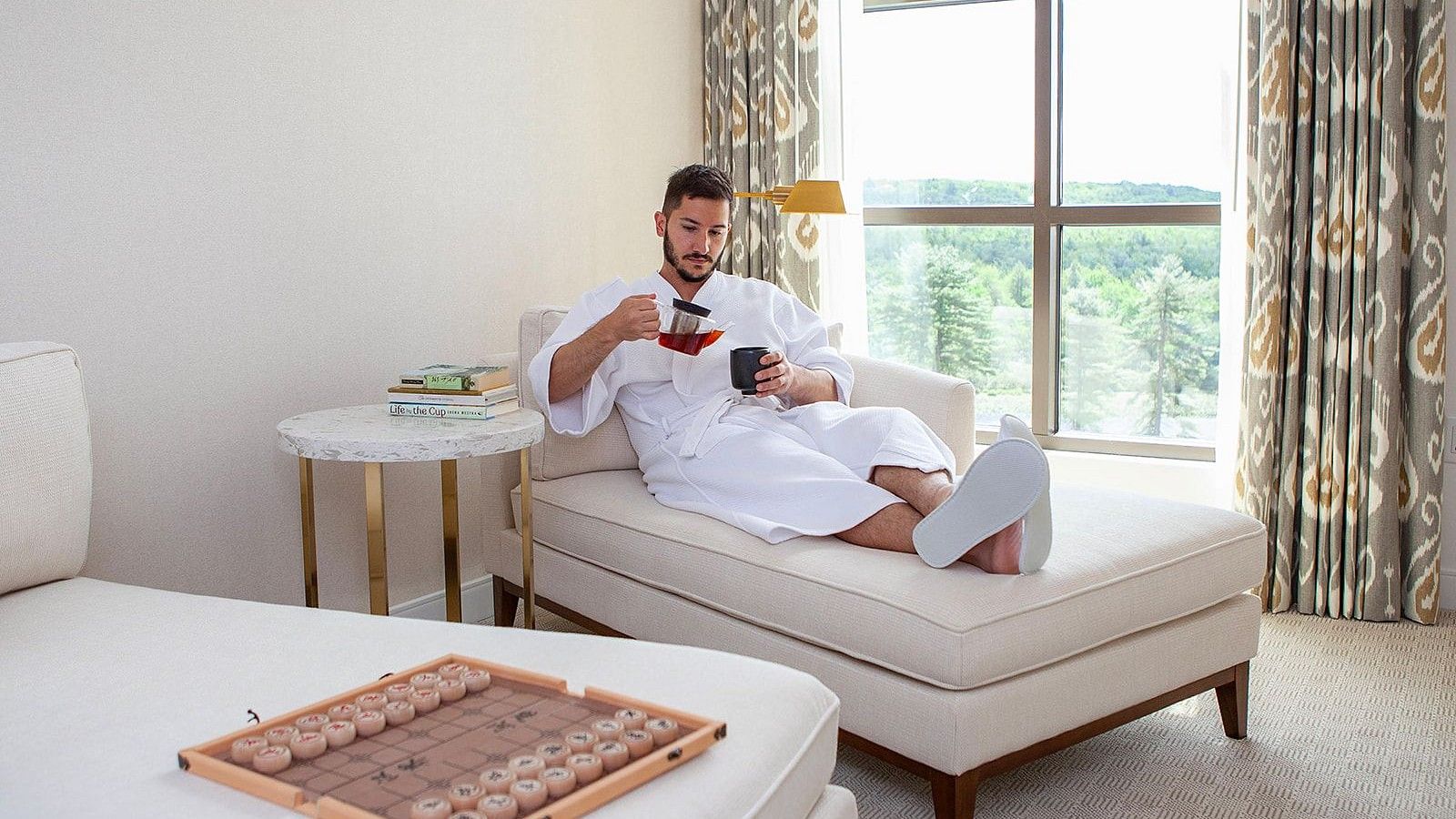 Man relaxing in a bright, elegant suite, wearing a bathrobe, sipping tea, with a board game nearby and large window view behind at YO1 Longevity & Health Resorts