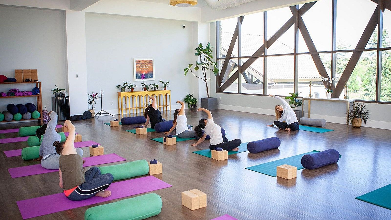 People practicing yoga at YO1 Longevity and Health Resorts in a bright studio with large windows, colorful mats, bolsters, and blocks, surrounded by plants and natural light.