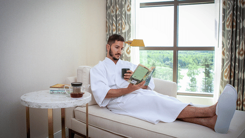 A man in a white robe and slippers relaxes on a chaise lounge, reading a book and drinking from a mug at YO1 Longevity & Health Resorts.
