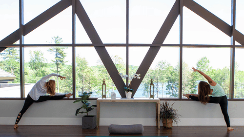 2 women in a bright studio with a large window stretching in a side-bending yoga pose with their arms raised at YO1 Longevity & Health Resorts