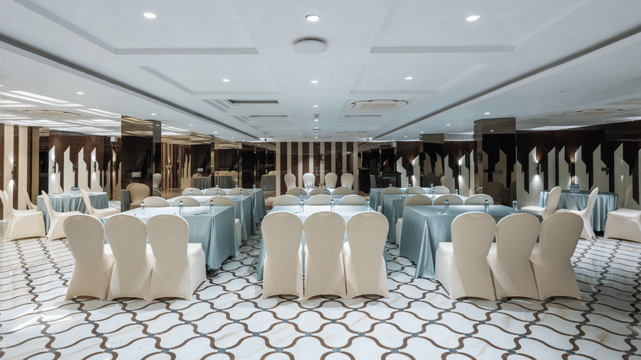 A banquet hall with white chairs arranged around round tables covered in blue cloths on a patterned floor | Lawrence Solitaire Resort, Nainital