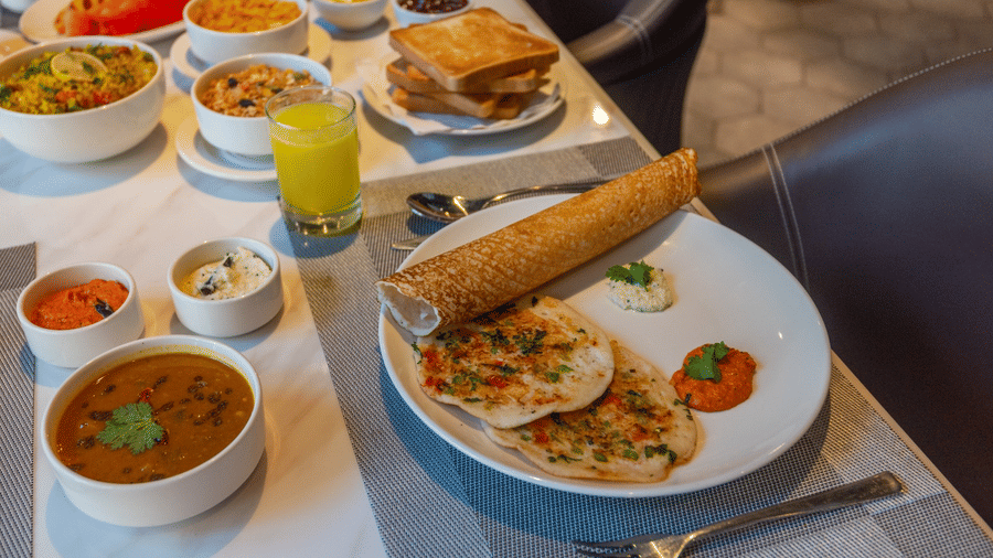 A breakfast spread on a table including a dosa, uttapam, various chutneys, sambar, toast, and orange juice | Lawrence Solitaire Resort, Nainital