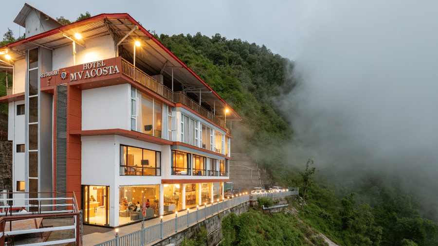 Modern hillside hotel building with illuminated interiors, surrounded by misty mountains - MV Acosta, Mussoorie 