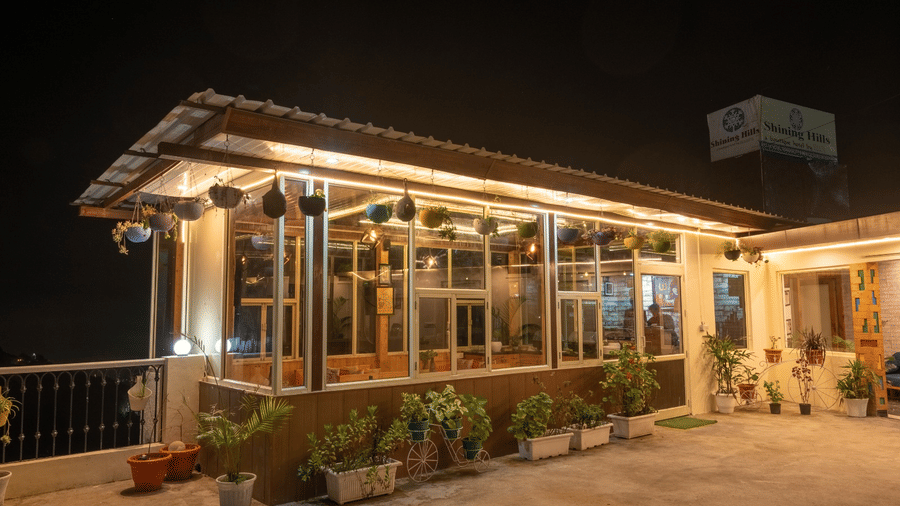 A brightly lit dining space of Shining Hills, Mussoorie, with a glass facade, a board with the hotel name on top and plants neatly arranged around the area.