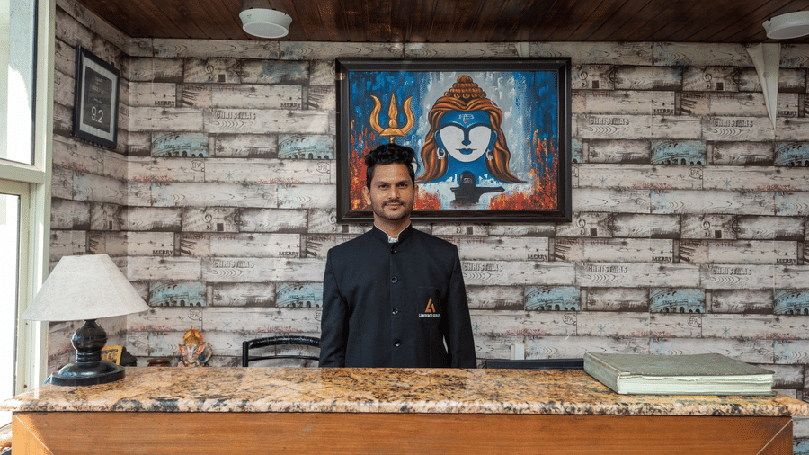 The hotel reception desk of Shining Hills, Mussoorie with a smiling staff, framed artwork, a lamp placed on the table and wooden interior.