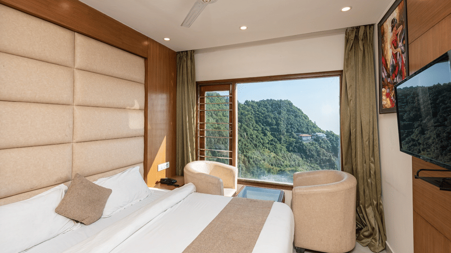 A hotel room at Shining Hills, Mussoorie with a large bed, padded walls, ceiling fan, two cushion chairs and a glass window showcasing the views of hills.