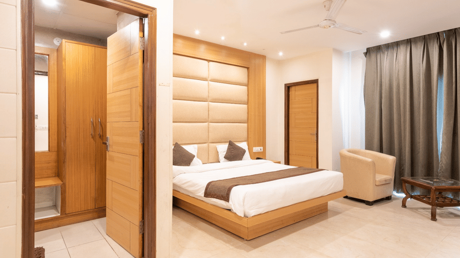 A hotel room at Shining Hills, Mussoorie with a large bed, padded headboard, ceiling fan, a door opening to another room, a cushion chair and bedside tables.