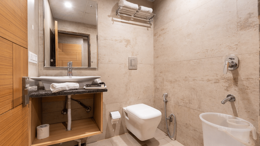 A bathroom at Shining Hills, Mussoorie with a toilet, wash basin with a towel kept aside, a mirror, and tiled walls.