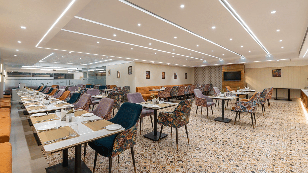 A spacious hotel restaurant with neatly arranged tables covered in beige placemats. The chairs are a mix of solid-coloured and floral-patterned upholstery. The ceiling features modern recessed lighting. The floor has an intricate geometric tile pattern. - 