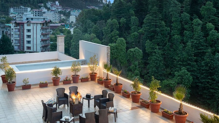 A rooftop terrace features a fire pit surrounded by chairs, potted plants, a shallow pool, and a view of buildings and lush greenery at Clarks Inn, Near Kainchi Dham.