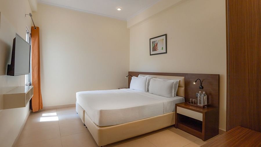 Bedroom featuring a double bed with white linens, a wall-mounted television, a bedside table with lamps and water, and a window with an orange curtain at Clarks Inn, Near Kainchi Dham