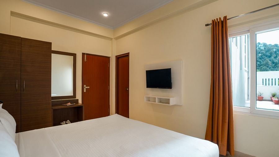 Room shows a double bed, a wardrobe, a dressing table with a mirror, a wall-mounted TV, and a window with an orange curtain at Clarks Inn, Near Kainchi Dham