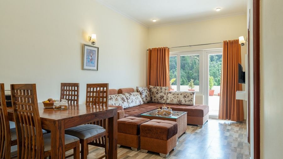 Living and dining space featuring a wooden dining set, a corner sofa with patterned cushions, pouffes, and patio doors with orange curtains at Clarks Inn, Near Kainchi Dham.