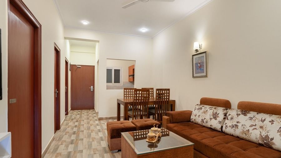 Living area featuring a brown corner sofa with floral cushions, a glass coffee table with pouffes, a dining table, and doorways at Clarks Inn, Near Kainchi Dham.