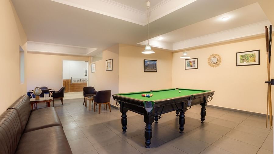 The game room at Clarks Inn, Near Kainchi Dham features a pool table with balls, a long brown sofa, armchairs around a small table, and framed pictures on the walls.